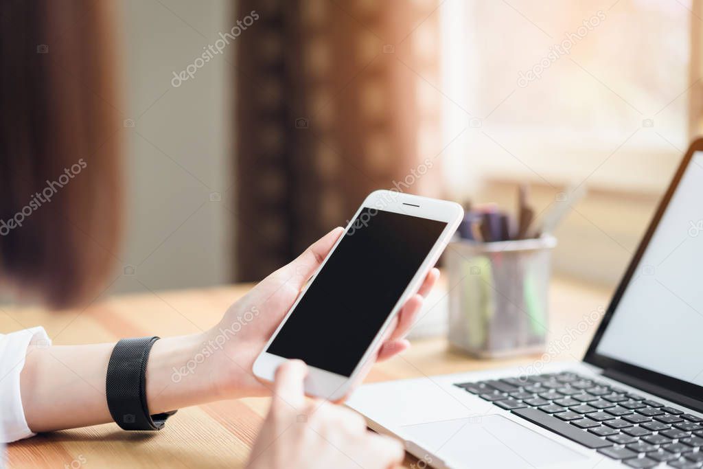 woman holding smartphone and using laptop on table in office room on windows with trees and nature background, for graphics display montage. Take your screen to put on advertising.