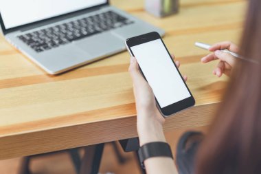 akıllı telefon tutarak ve montaj grafikleri görüntülemek için tablo ile ağaçlar ve doğa arka plan, windows üzerinde oda ofis dizüstü bilgisayar kullanarak bir kadın. Senin ekran reklam koymak için almak.