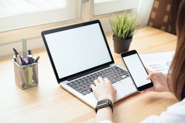 akıllı telefon tutarak ve montaj grafikleri görüntülemek için tablo ile ağaçlar ve doğa arka plan, windows üzerinde oda ofis dizüstü bilgisayar kullanarak bir kadın. Senin ekran reklam koymak için almak.