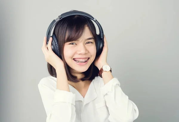 在白色休闲礼服的亚洲女孩听音乐从黑色耳机 在一个舒适和良好的心情 在灰色的背景给一个柔和的光 — 图库照片