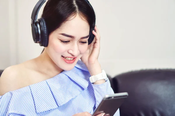 Asian girl in blue casual dress listening to music from black headphones. In a comfortable and good mood, In the living room on the sofa.