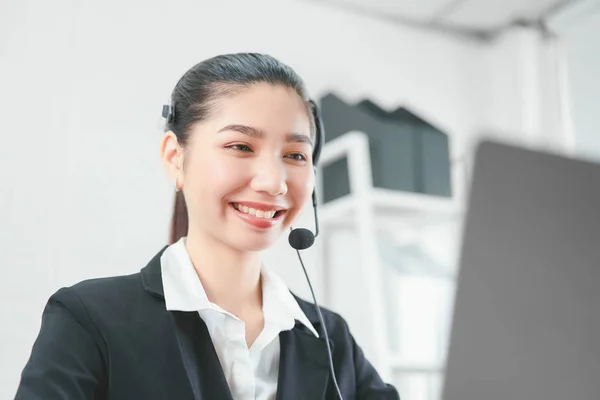 Smiling Asian woman consultant wearing microphone headset of customer support phone operator at workplace.