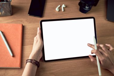 Boş ekranlı tablet kullanan ve dijital kalem tutan eller, kameralı akıllı telefon ve ahşap masadaki aksesuarlar..