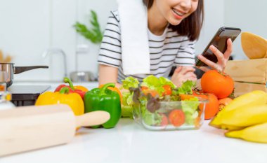 Elinde akıllı telefon ve domatesli salata kasesi ve evdeki masada yeşil yapraklı sebzeler olan bir kadın, reklamınızı alın..