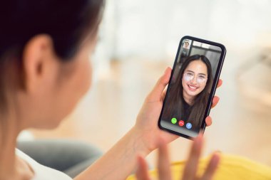 Genç gülümseyen Asyalı kadın internetten video görüşmesi yapıyor arkadaşlarıyla evden akıllı telefona konuşuyor..