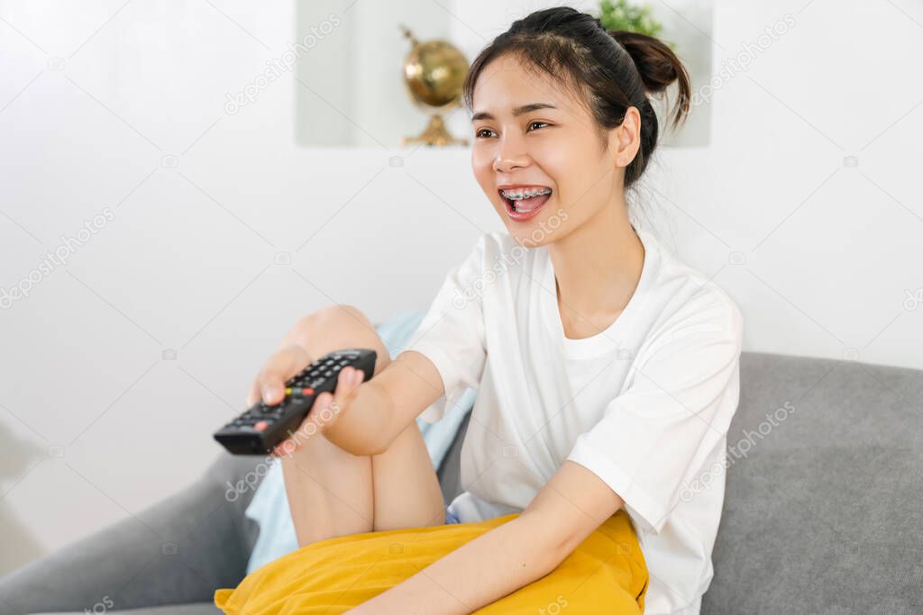 Young Asian woman sitting at the sofa in the house and holding the TV remote to watch movies on a relaxing day.