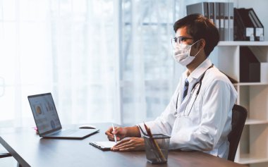 Maskeli doktor bir masada oturuyor ve hastanın durumunu nasıl tedavi edeceğini araştırıyor..