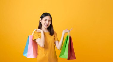 Genç gülümseyen Asyalı kadın açık sarı arka planda çok renkli alışveriş torbaları tutuyor..