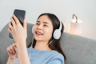 Mutlu gülümseyen Asyalı kadın beyaz kulaklıklardan müzik dinliyor ve evde tatilde akıllı telefonlara dokunuyor..