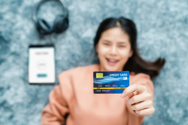 笑着躺在地板上的亚洲女人 并在数码平板电脑上拿着装有移动钱包应用程序的信用卡 图库照片