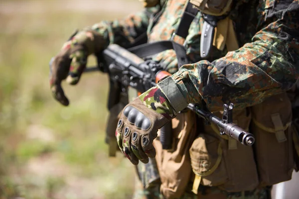 A soldier in gloves and green camouflage holds his hands on a machine gun that hangs on his neck. AK47 assault rifle close-up