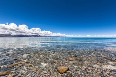 Büyüleyici manzara Namtso Gölü (Nam Gölü, Tengri Nor) Batı Nyenchen Tanglha Dağları Qing Zang Platosu 'nda, güneşli yaz günü mavi gökyüzü, temiz su ve taş, Tibet, Çin.