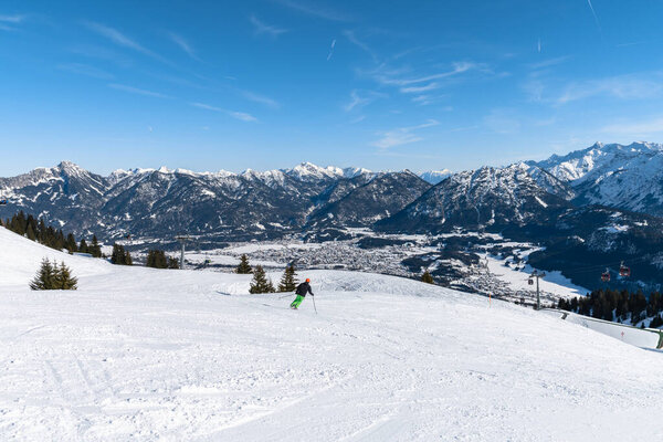 Panorama view snow covered Austrian Alps mountain range in winter sunny day clear blue sky above Reutte Hahnenkamm ski resort, ski slope  and people skiing in foreground, Tyrol Tirol Austria