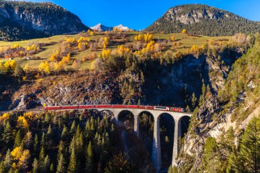 Rhaetian Demiryolu treni ünlü Landwasser Viyadüğü 'nde güneşli bir sonbahar gününde renkli ağaçların olduğu tünele doğru ilerliyor.
