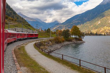 Sonbaharda Lago di Poschiavo gölünün kenarında mavi gökyüzü bulutuyla koşan kırmızı Rhaetian treninin güzel manzarası, demiryolu hattı Bernina Express, Grisonlar Kanonu, İsviçre