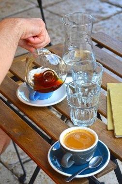 Erkek eli filtre kahve ve espresso Lungo 'yu mavi bardaklara dolduruyor. Dışarıdaki ahşap masada bir sürahi suyla birlikte.