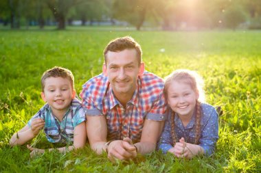 Parkta neşeli aile, baba ve ona iki güzel çocuk, erkek ve kız onlar kameraya bakarken çim üzerinde yatıyor. Arka planda gün batımında. Mutlu sevgi dolu aile kavramı