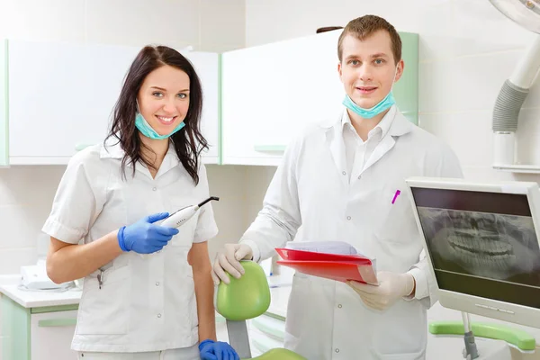 患者のための椅子の近くに歯科医院のキャビネットに立って笑顔の若い歯科医とアシスタント — ストック写真
