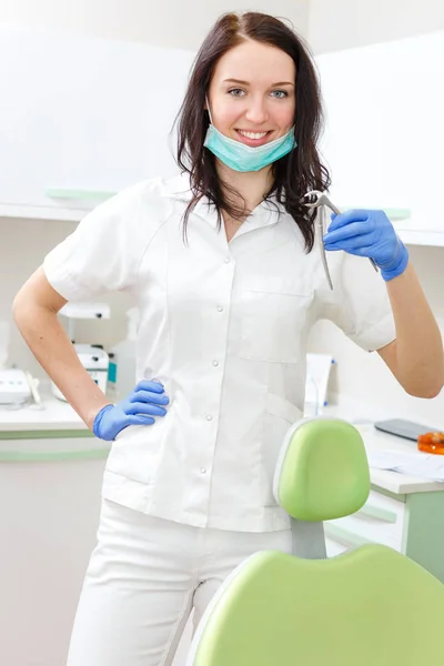 年轻美丽的女牙医笑着高高兴兴地站在牙椅前的办公室里 拿着牙具 — 图库照片