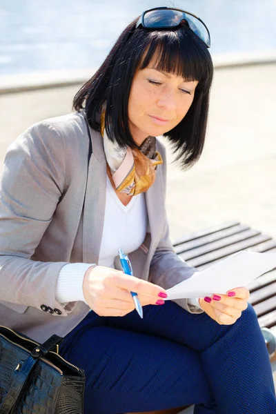 Stylish brunette pretty woman writing a letter and thinking with Pen in hand, sitting on a park bench, sunny day, good weather, city street, human emotion, facial expression