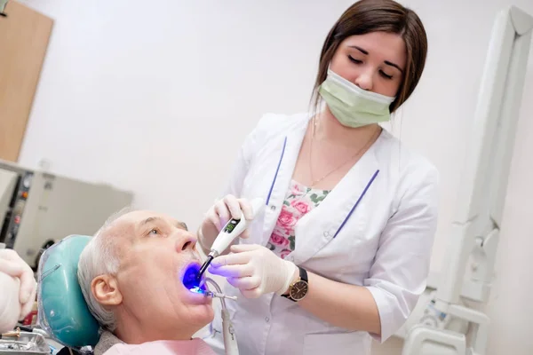 シニア男性患者の口腔内の歯科重合ランプで働く歯科医女性 高齢者の歯科治療歯科用デバイスを保持する歯科医 高齢者の歯科医療 — ストック写真
