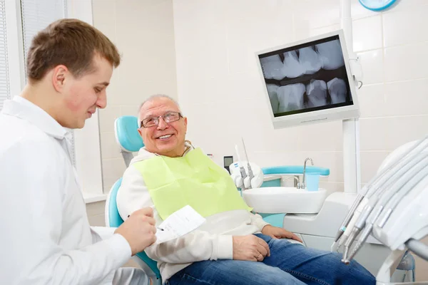 Happy and smile senior man 70-75 years old with white teeth on review of a dentist, sitting in chair dental clinic. Looking on young dentist doctor. Dental care for older people. Health care concept