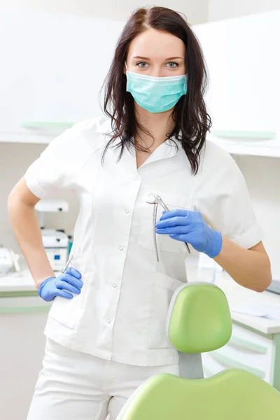 年轻漂亮的戴着防护面具的女牙医站在牙医椅子前的办公室里 手里拿着牙科工具 — 图库照片