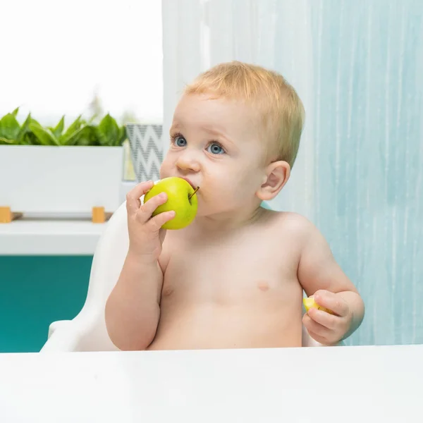 Menino loiro com olhos blu comer maçã na cozinha perto da janela. espaço de cópia — Fotografia de Stock