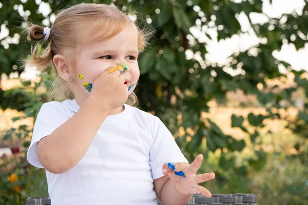 toddler girl touches her nose with a finger covered in paint.