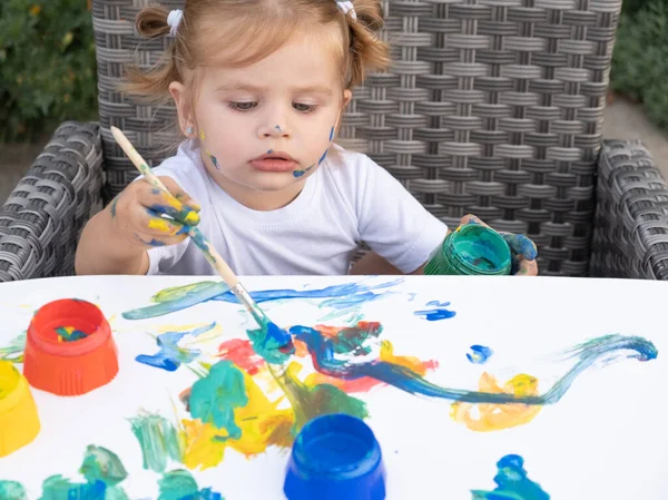 Милый очаровательный белый маленький блондинка девочка рисует с краской и щеткой за дворовым столом. — стоковое фото