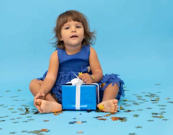 Petite fille brune en robe bleue assis détache boîte cadeau sur fond bleu. — Photo