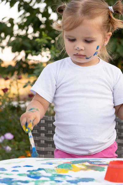 Маленький ребенок рисует краской и кистью. Cute little girl painting picture in garden, outdoors at home in backyard. — стоковое фото