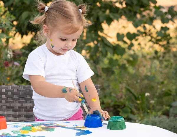 Маленький ребенок рисует краской и кистью. Cute little girl painting picture in garden, outdoors at home in backyard. — стоковое фото