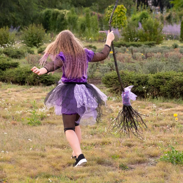 Młoda dziewczyna w stroju czarownicy halloween fioletowy trzymając miotłę i biegając po trawniku. — Zdjęcie stockowe