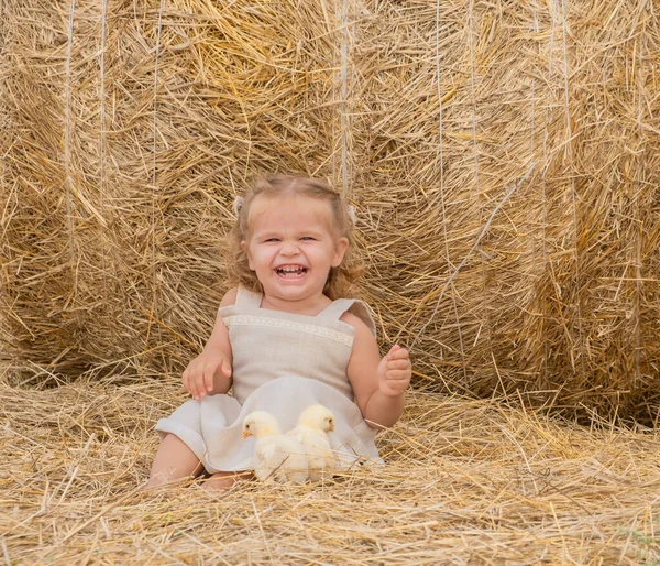 Счастливый ребенок в льняном платье смеется с пушистым ребенком цыплят улыбается сидя на сене. — стоковое фото