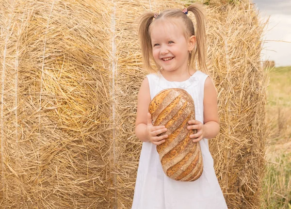 Beyaz elbiseli küçük sarışın kız tarlada ekmek tutuyor. Çevre dostu tarım ürünleri.