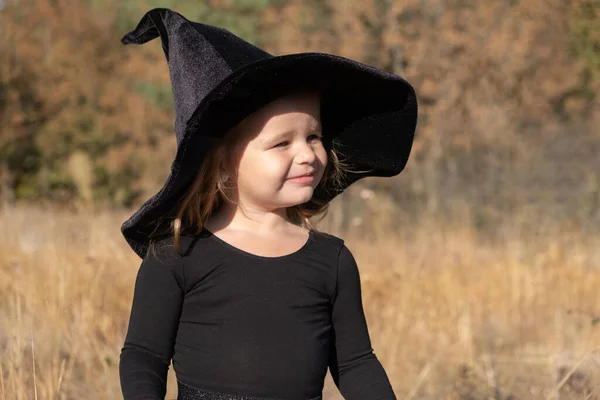 Χαριτωμένο κοριτσάκι σε μαύρο καπέλο μάγισσα στη φύση. — Φωτογραφία Αρχείου