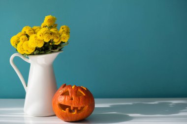 Masanın üzerinde sarı çiçekli beyaz bir vazo ve balkabağı var..