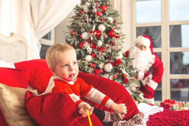 Küçük üzgün çocuk şenlikli bir yatak odasında bir yatak üzerinde oturuyor. Gerçek Noel Baba ondan Noel ağacın arkasına saklanıyor.