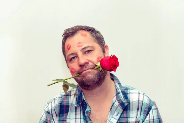 Bearded mature man with a red rose in his mouth. There are traces of lipstick on his face.