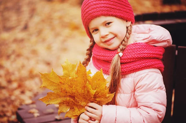 葉の花束を持つ子少女の笑顔 暖かいニット帽子とスカーフで公園のベンチに座って 晴れた日の屋外散歩を楽しむ秋の肖像画 — ストック写真