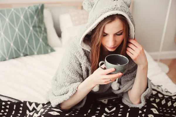 若くてきれいな女性のベッドの家でのんびりおかしい灰色のパーカーを着ています 紅茶やコーヒーのカップと怠惰な週末の朝の低温 — ストック写真