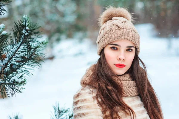 winter portrait of beautiful young woman in fur coat and knitted hat walking in snowy park or forest.