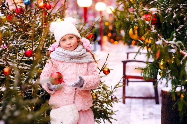 雪に覆われた冬を歩く幸せな子女の子のクリスマスの肖像画には 背景に木が飾られています 市の年末年始 — ストック写真