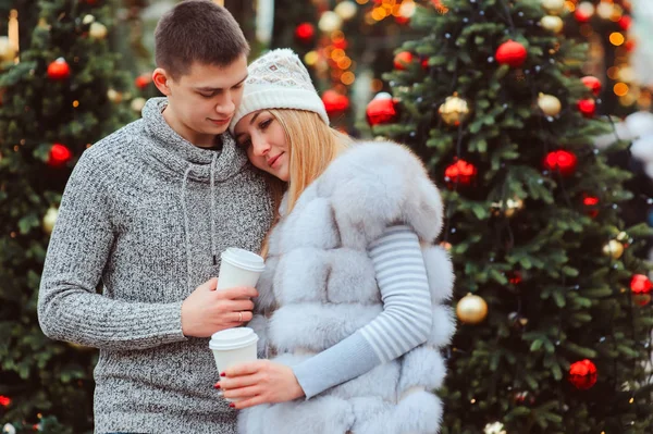 christmas portrait of happy couple with hot mulled wine or tea walking on city streets decorated for holidays, christmas trees with lights and toys on background
