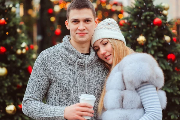 christmas portrait of happy couple with hot mulled wine or tea walking on city streets decorated for holidays, christmas trees with lights and toys on background