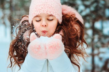 Beyaz önlüklü, şapkalı ve pembe eldivenli mutlu bir kızın kış portresi. Karlı kış ormanlarında dışarıda oynuyor ve kar üflüyor. Mutlu çocukluklar ve aktif kış tatili konsepti