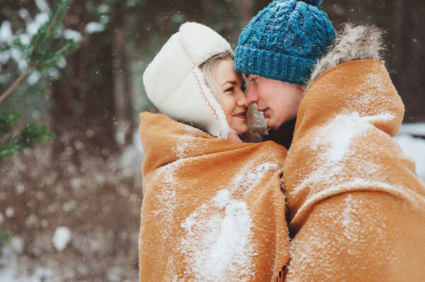 happy young loving couple walking in snowy winter forest, covered with oversize scarf and hug. Active winter vacations outdoor.
