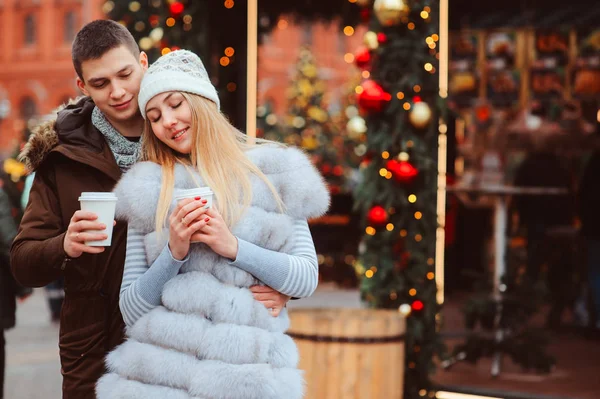 christmas portrait of happy couple with hot mulled wine or tea walking on city streets decorated for holidays, christmas trees with lights and toys on background