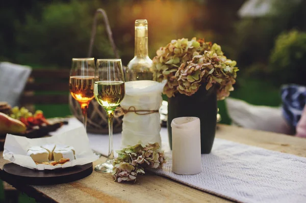 evening summer garden party with wine, cheese and fruits on wooden table decorated with candles and flowers